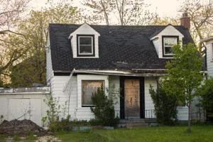 Cape Cod cottage before — boarded windows, damaged roof, overgrown and abandoned